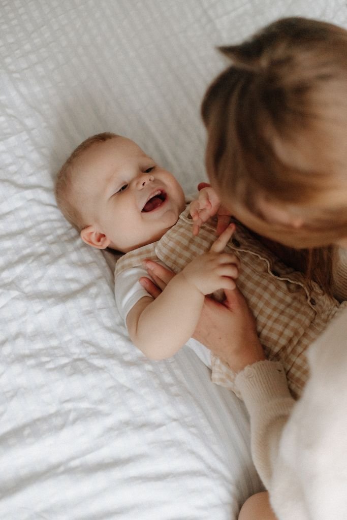 A mother playfully interacts with her laughing baby on a cozy bed.